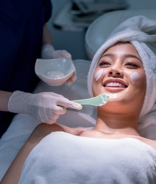 Wide shot hands hold paddle apply beauty cream on face of beautiful woman during process in beauty clinic or salon.